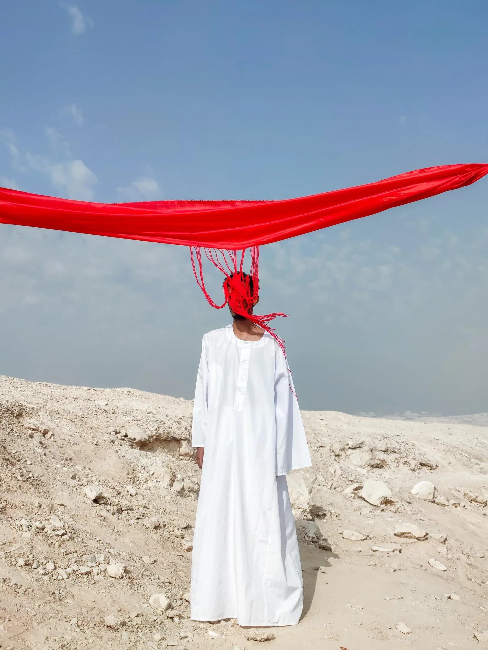 Figure in white robe standing on sandy terrain beneath a large red fabric suspended overhead against pale sky.