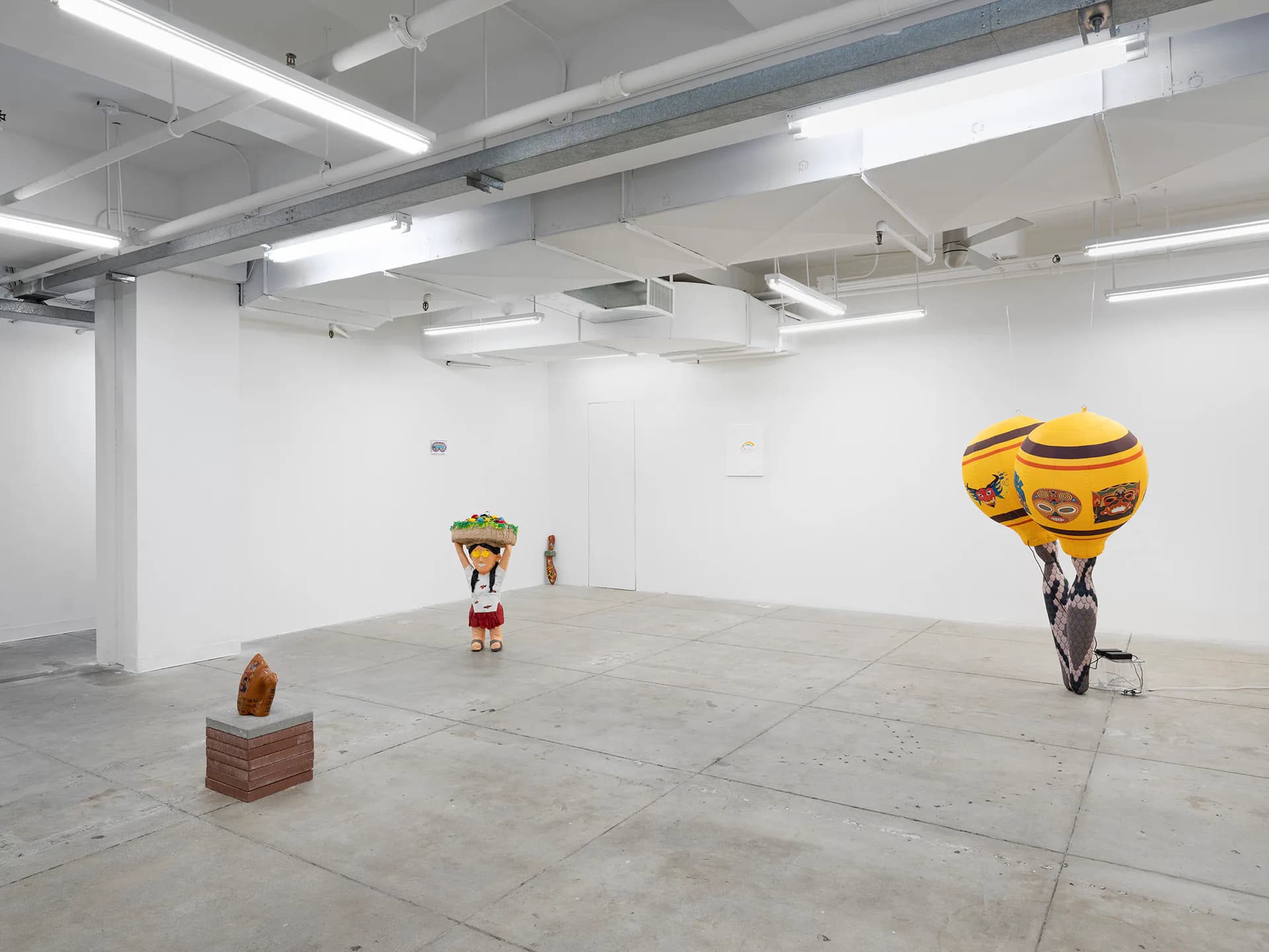 Mama Projects gallery interior: white-walled industrial space with polished concrete floor, fluorescent ceiling lights, displaying sculptural works including yellow and brown forms.