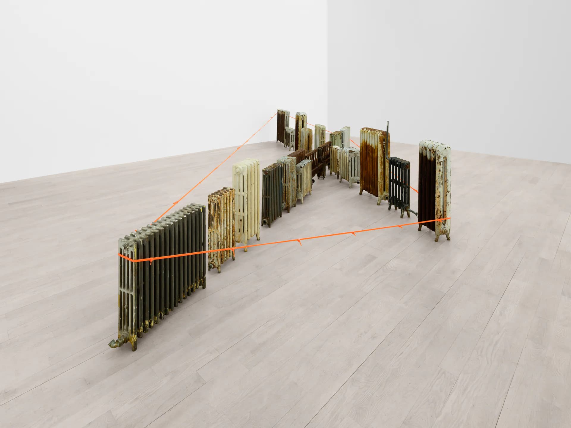 Installation artwork of stacked brass tubes and metal rods arranged in clusters with orange line connecting forms across light gallery floor.