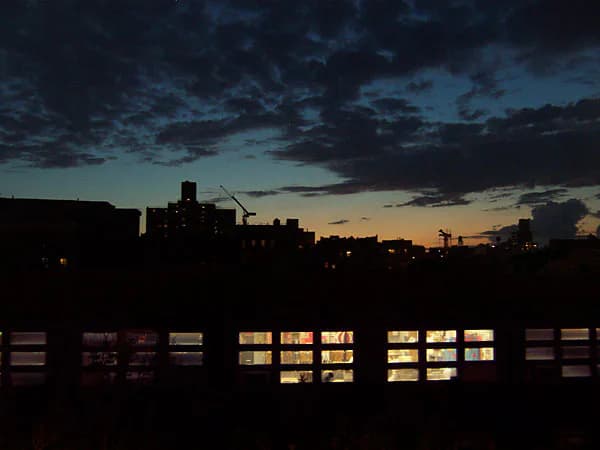 Twilight cityscape of Manhattan silhouette against dusky sky with lit windows in foreground building, mixed-media photograph capturing urban evening atmosphere.