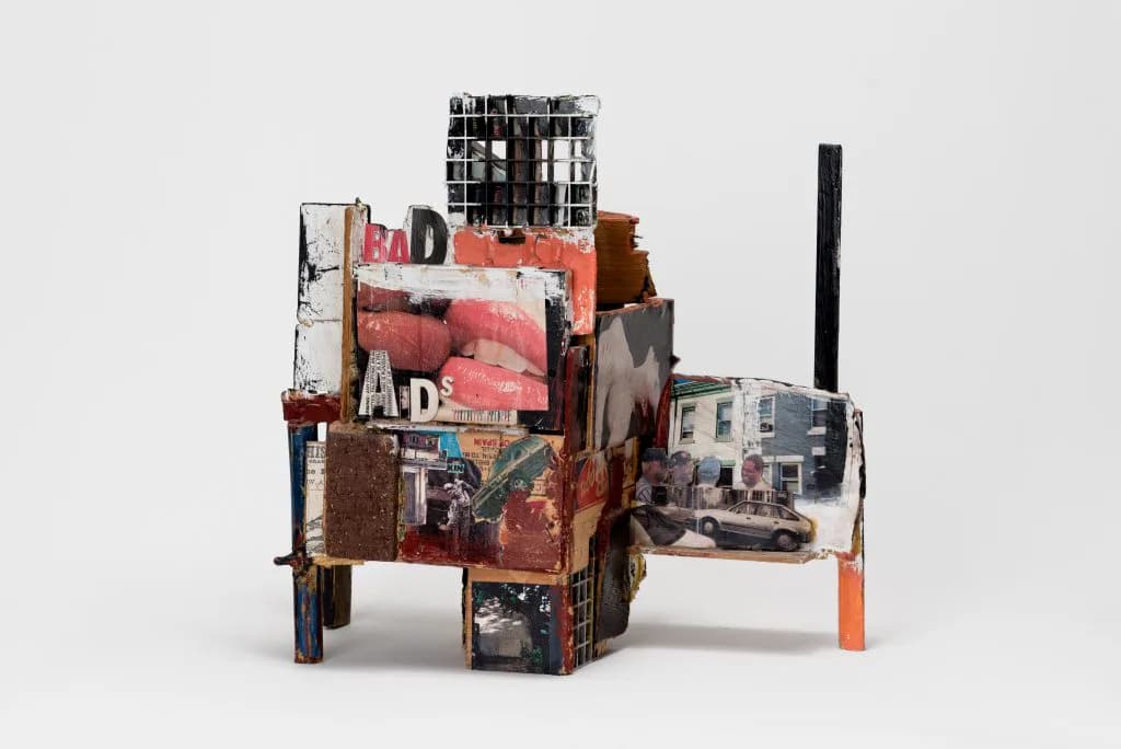 Mixed-media assemblage sculpture featuring stacked red, brown, and weathered metal/wood forms with collaged text and images, mounted on wooden supports.