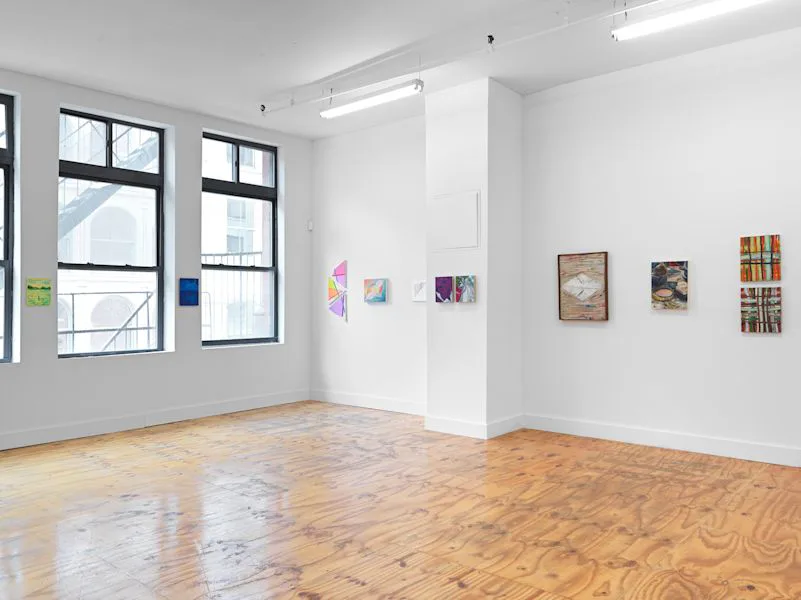 Gallery interior with central white dividing wall, large windows to the left, wood floors, and small colorful artworks hung on white walls.