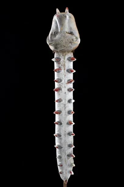 White sculptural form with animal-like head and elongated segmented body with red circular details, photographed against black background.