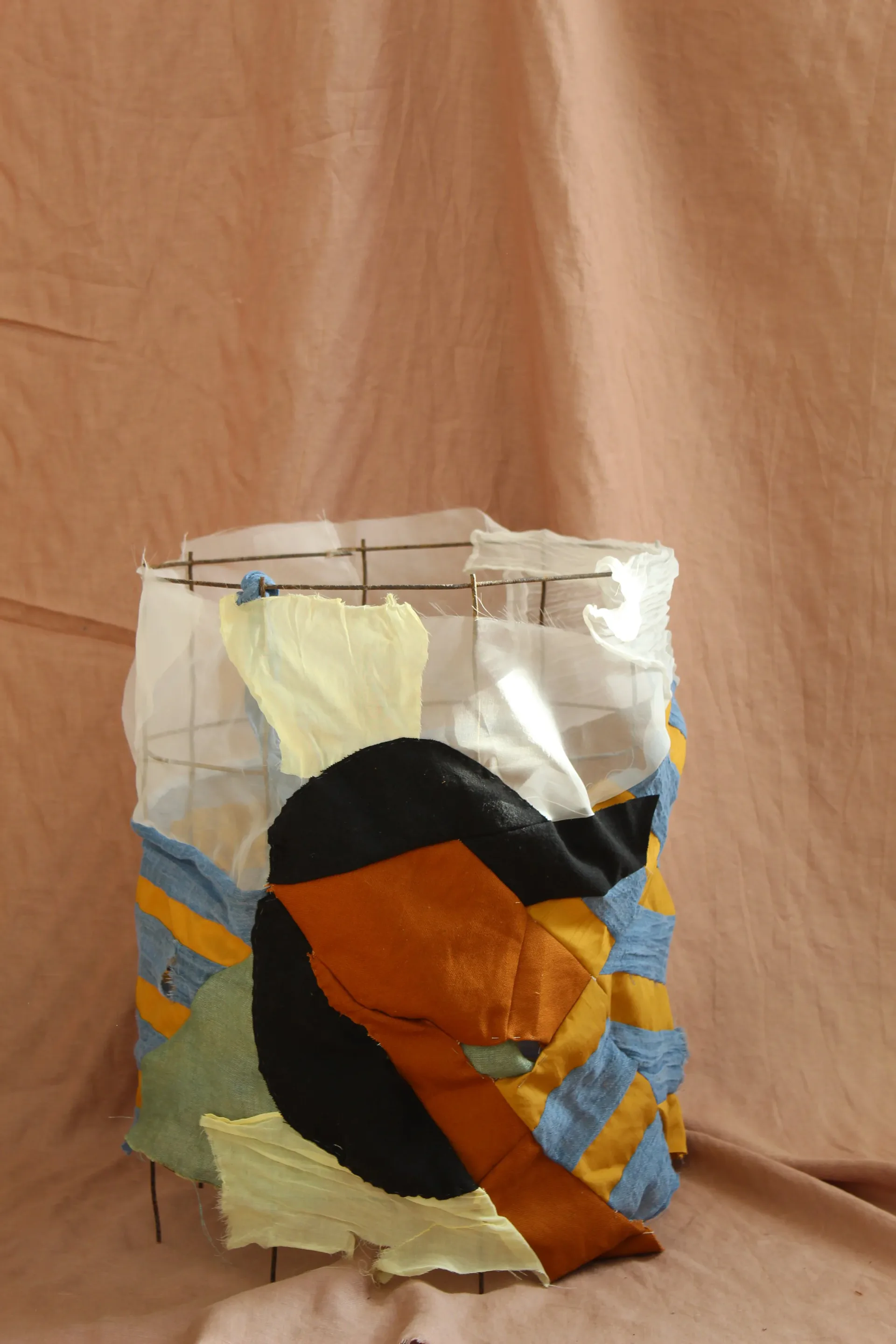 Cylindrical fabric sculpture with mixed colored panels including yellow, orange, black, and white, suspended on metal frame with clothespins.