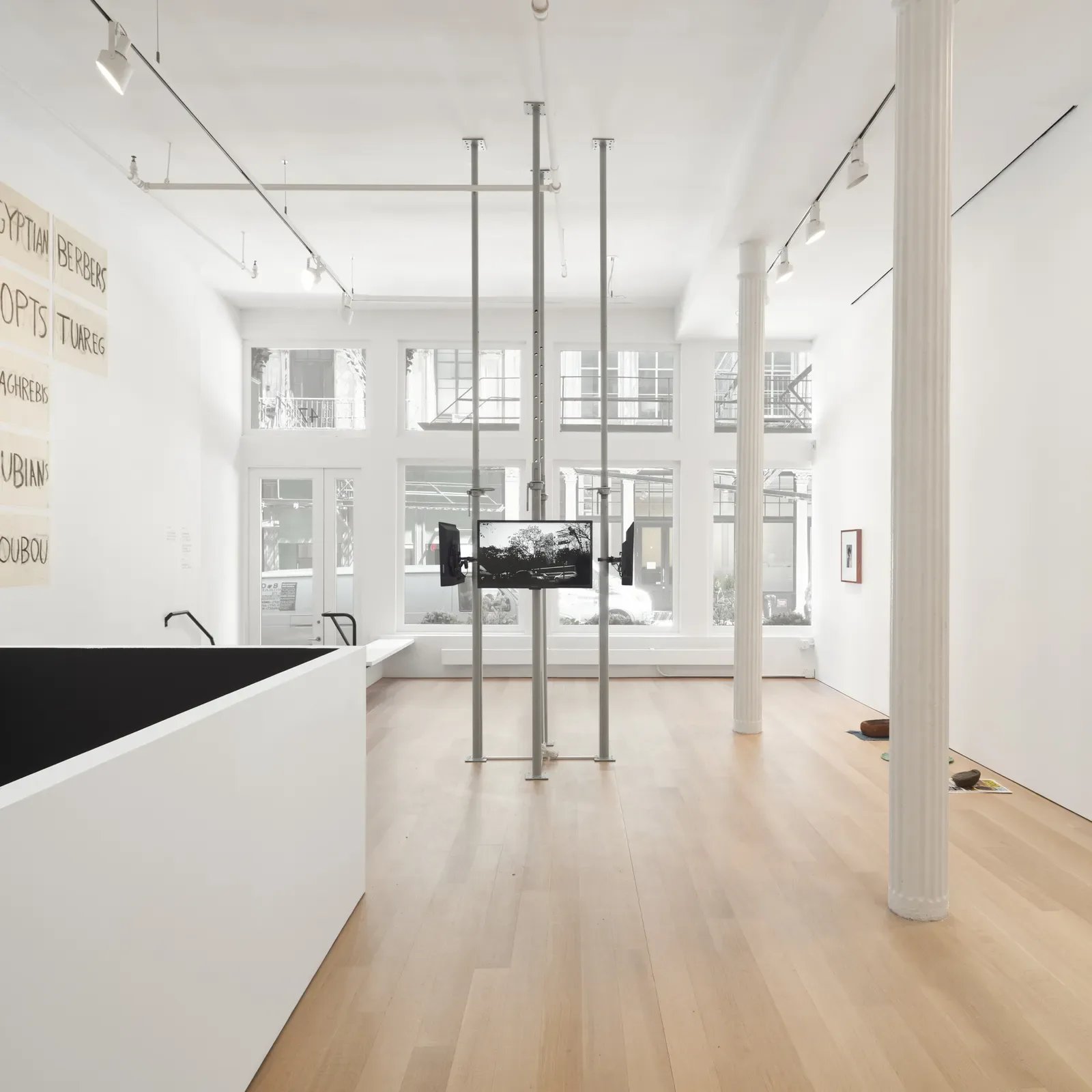 Bright gallery interior with white walls, wood floors, classical columns, and large windows overlooking the street