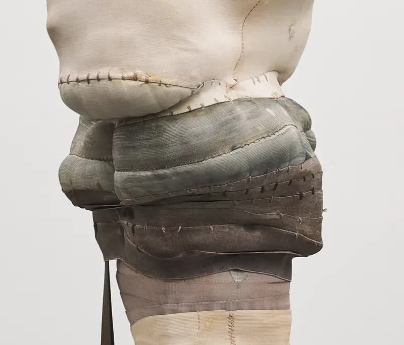 Sculptural fabric work showing layered textiles in gray and neutral tones wrapped and stitched together, displayed against white background.