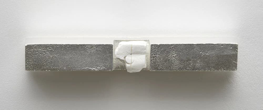 Minimalist sculptural work featuring two concrete blocks with white geometric form nestled between them on a neutral surface.