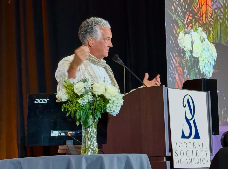 Man with gray hair speaking at podium with white flowers, wearing light-colored shirt, with projection screen visible behind him and Portrait Society of America logo on podium.