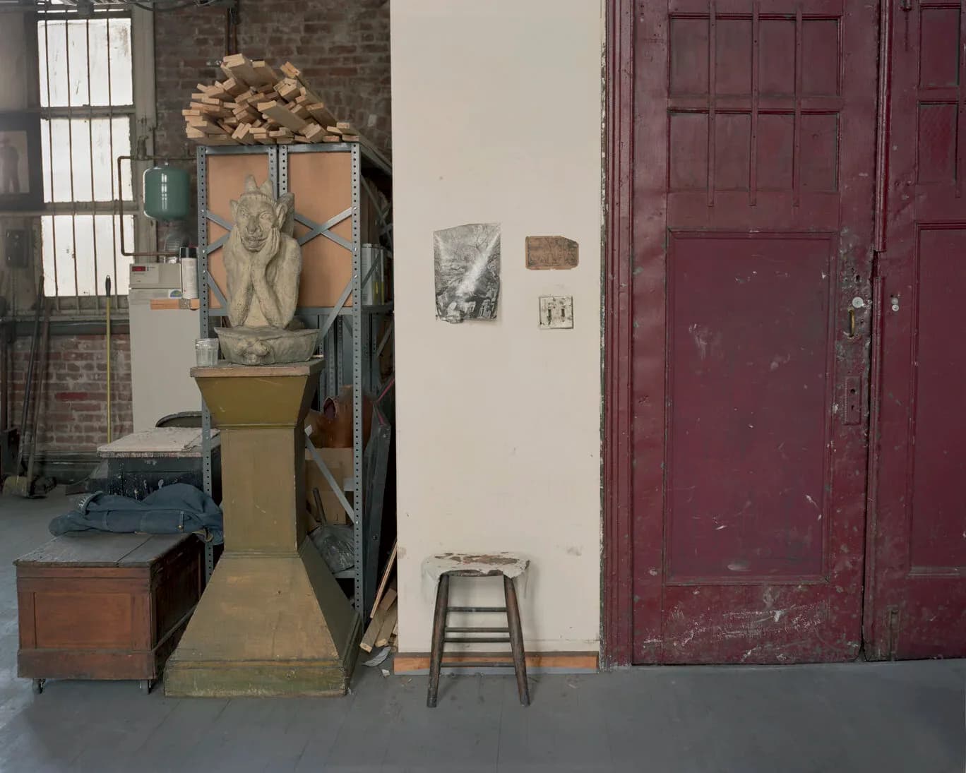 Gallery interior with industrial brick walls, sculptural pedestals displaying objects, a burgundy wooden door, metal stool, and window with metal bars.