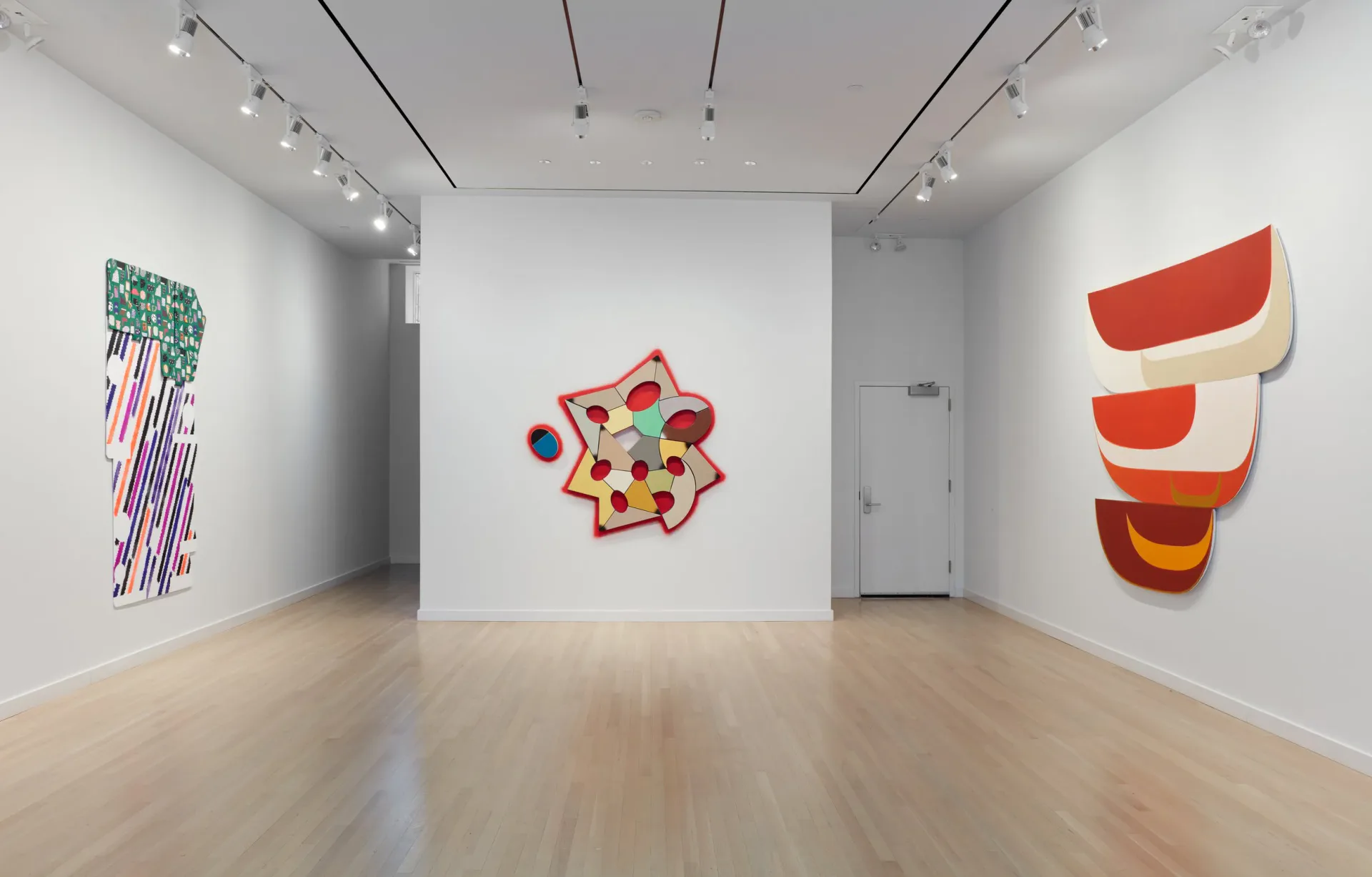 Bright gallery interior with track lighting, wooden floor, and three colorful abstract artworks displayed on white walls.