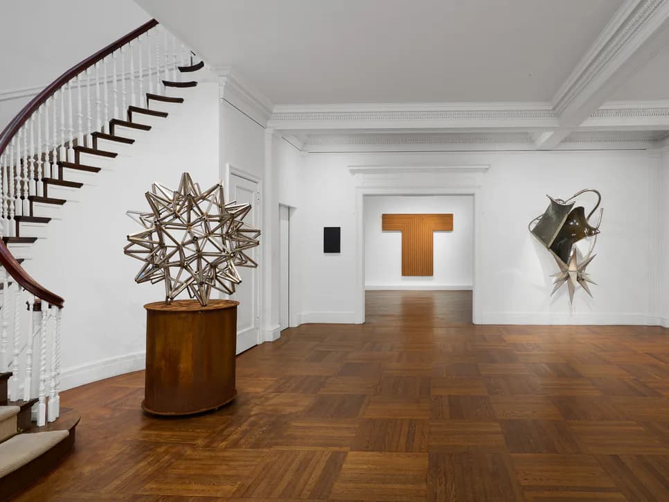 Gallery interior with wooden floor, curved staircase, and sculptural works including geometric star and bronze forms on display.