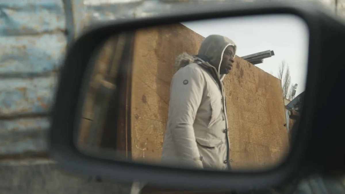 Person in light-colored clothing viewed through a vehicle's side mirror against a tan/brown wall background.