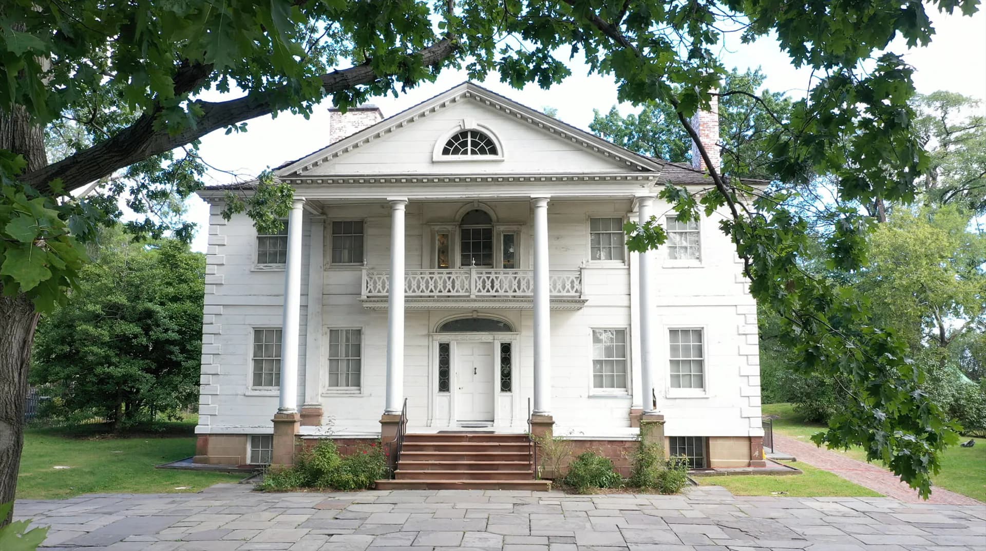 White neoclassical mansion with columned portico, brick foundation, and manicured grounds framed by large trees.