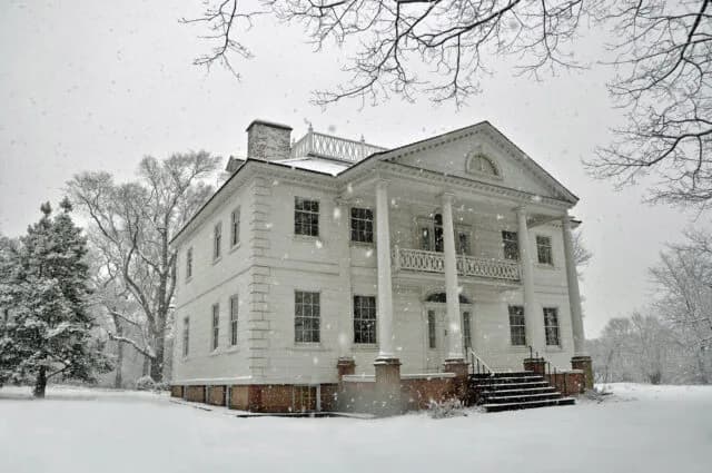 Morris-Jumel Mansion exterior in winter snow, white clapboard structure with columned facade and bare trees.