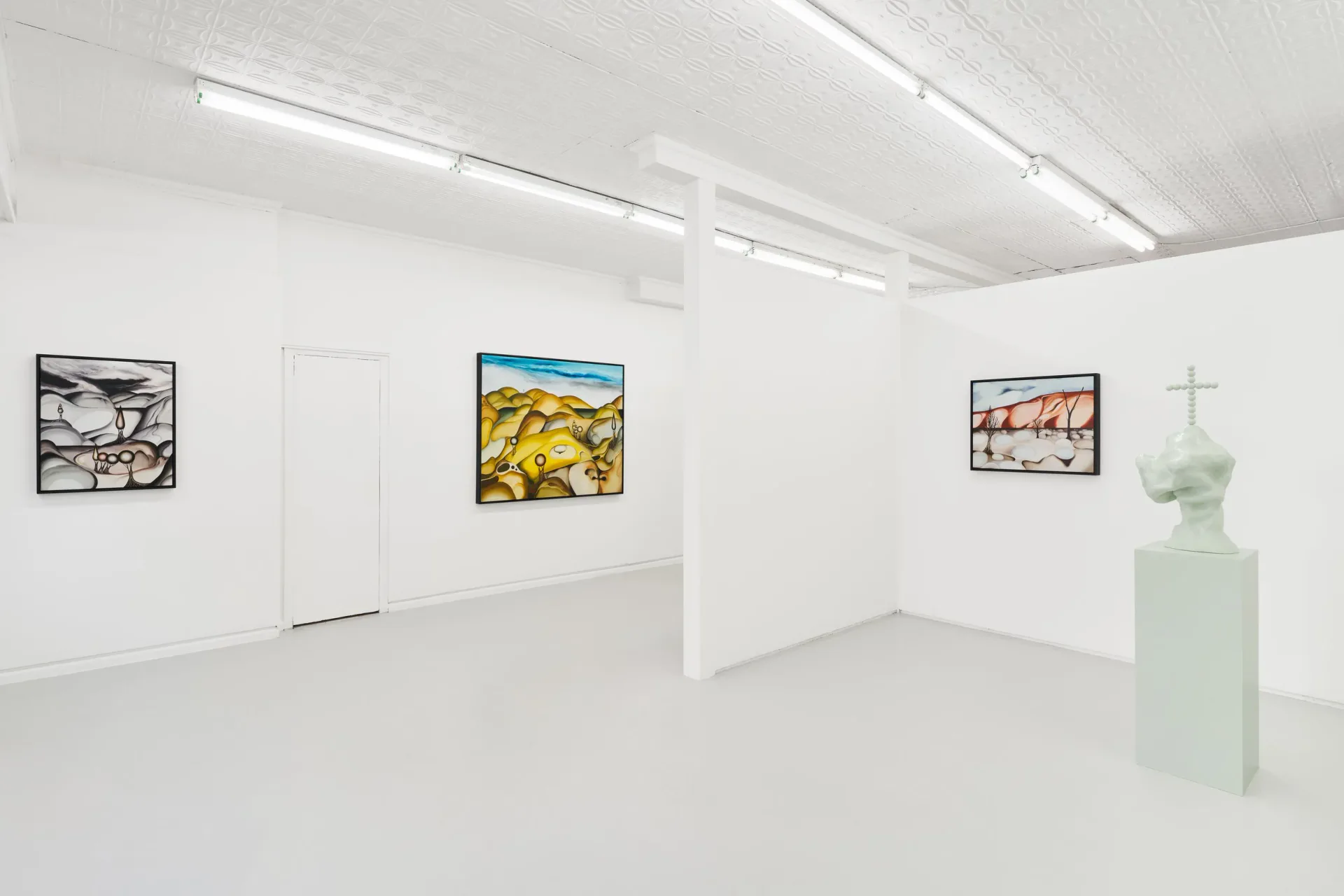 Bright gallery with white walls displaying three small framed images and a pale green sculptural bust on a pedestal in the corner.