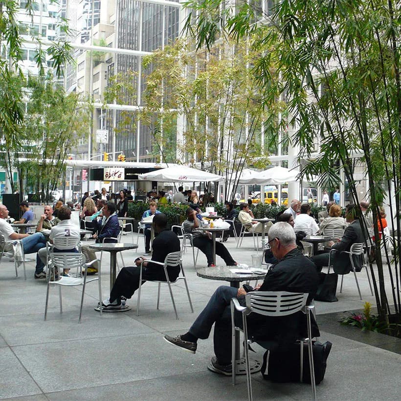 Urban courtyard plaza with seated visitors, bamboo trees, modernist buildings, and white tent structure overhead.