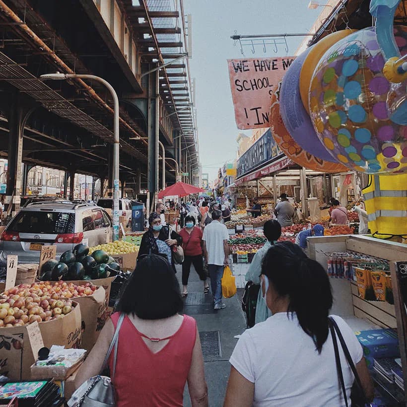 Busy outdoor market under elevated highway structure with produce stalls, vendors, and crowds of shoppers.