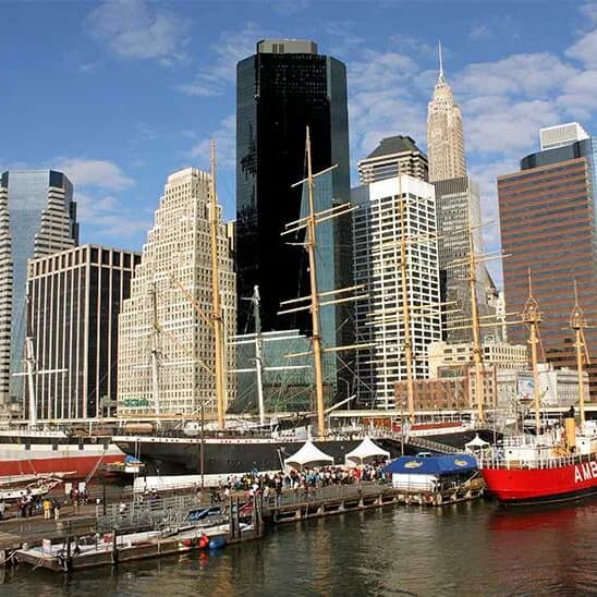 Manhattan waterfront skyline with iconic skyscrapers, including Empire State Building, viewed from pier with boats and crowds.