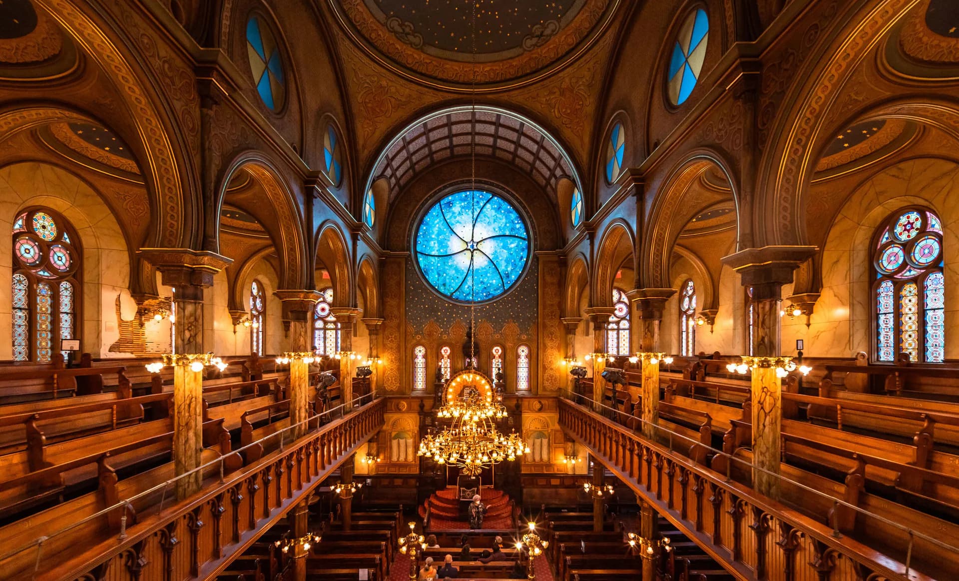 Ornate sanctuary interior at the Museum at Eldridge Street, viewed from the balcony with wooden pews, carved columns, and a large round stained-glass rose window in blue tones.