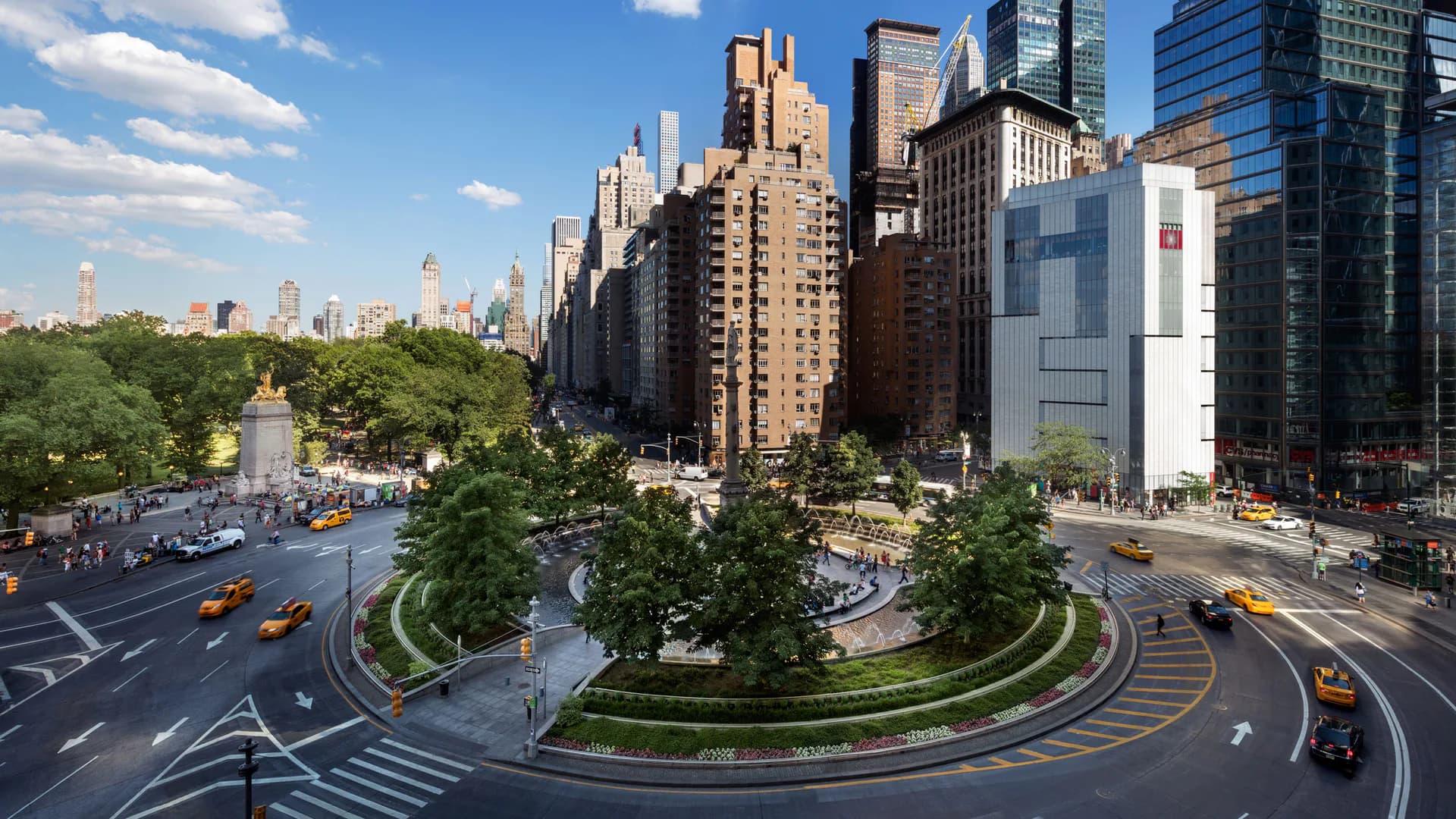 Aerial view of Columbus Circle in Manhattan showing a circular plaza with landscaping, surrounding tall buildings, yellow taxis, and pedestrians on streets.