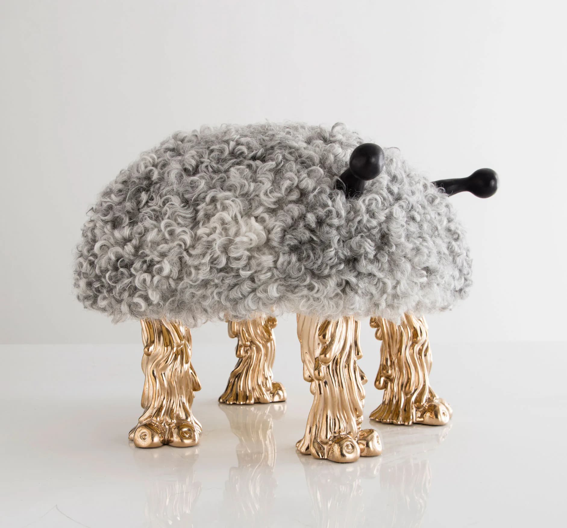 Sculptural artwork of a curly-haired creature with black antennae supported by four golden cast legs on a white surface.