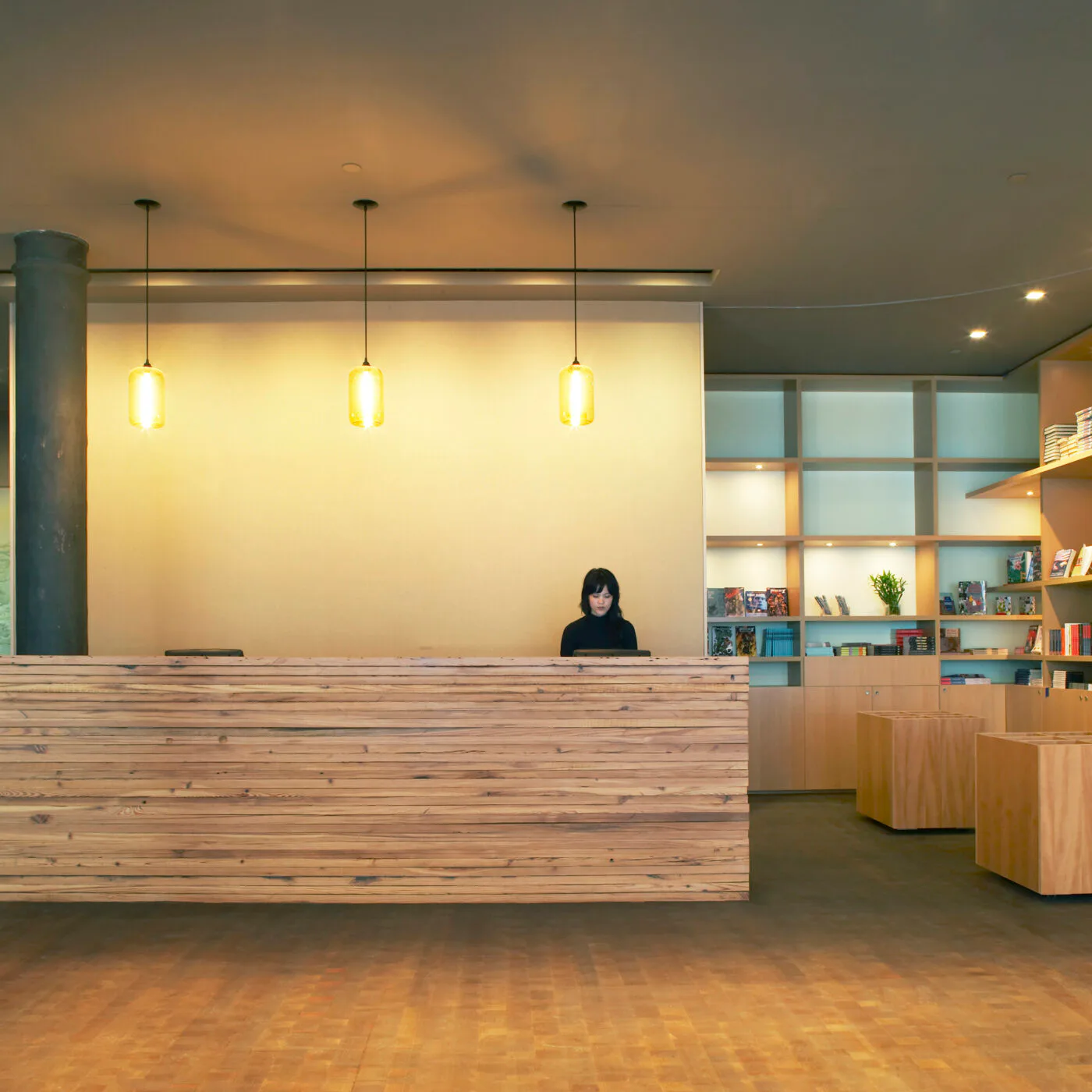 Modern gallery reception desk with wooden front and pendant lighting, shelving with turquoise backing visible behind, warm ambient lighting.