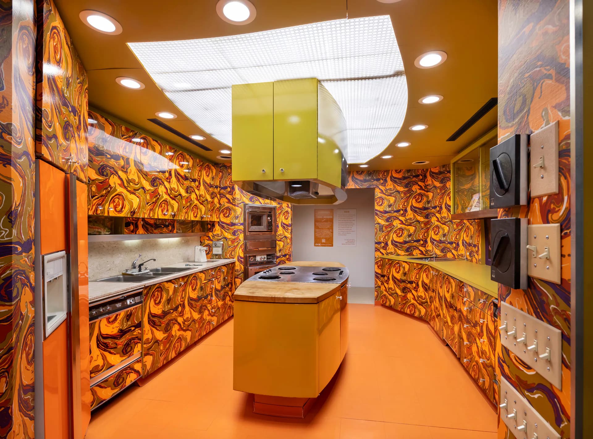 Interactive kitchen exhibition space at Museum of Food and Drink with orange walls, decorative patterns, cooking stations, and illuminated ceiling.