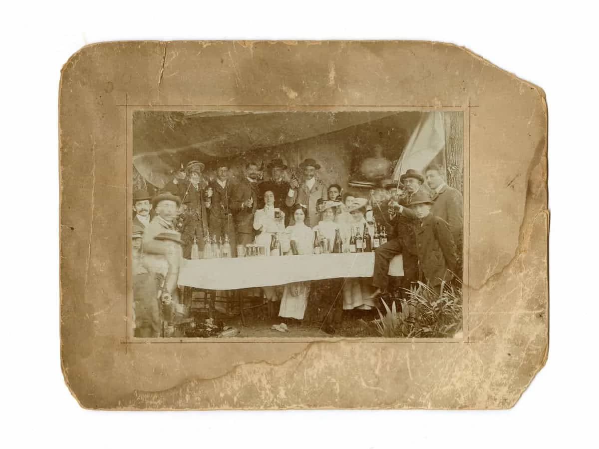 Sepia-toned vintage photograph mounted on aged paper showing large group of people gathered around a table indoors.