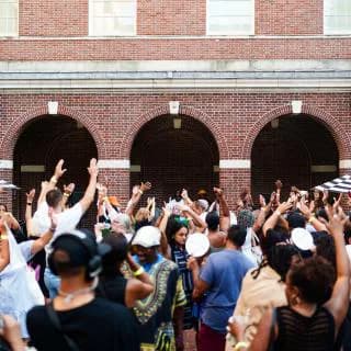 Outdoor crowd gathered with raised hands in front of a brick building with three dark arched entrances and light fixtures.