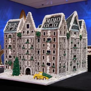 Intricate gingerbread building model featuring three connected ornate structures with detailed architectural elements and a small yellow toy vehicle.