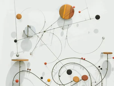 Kinetic mobile sculpture with intersecting wire frames, wooden shelves, and colorful spheres in orange, black, and gray suspended against a white wall.