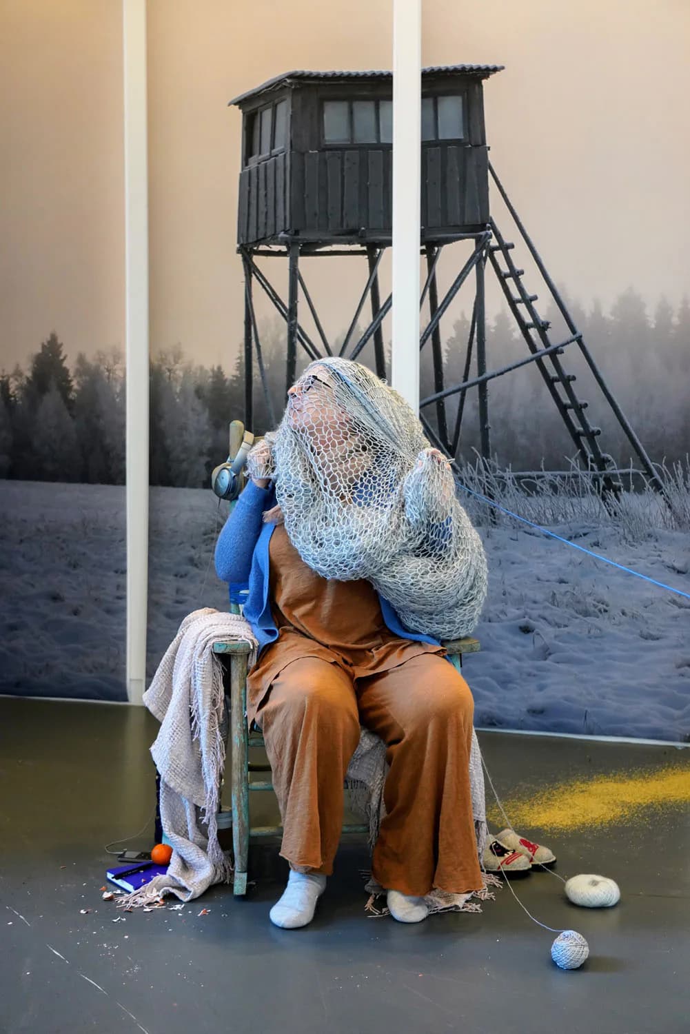 Figure wearing tan clothing and large crocheted gray head covering seated before landscape photograph and elevated wooden structure; mixed-media installation.