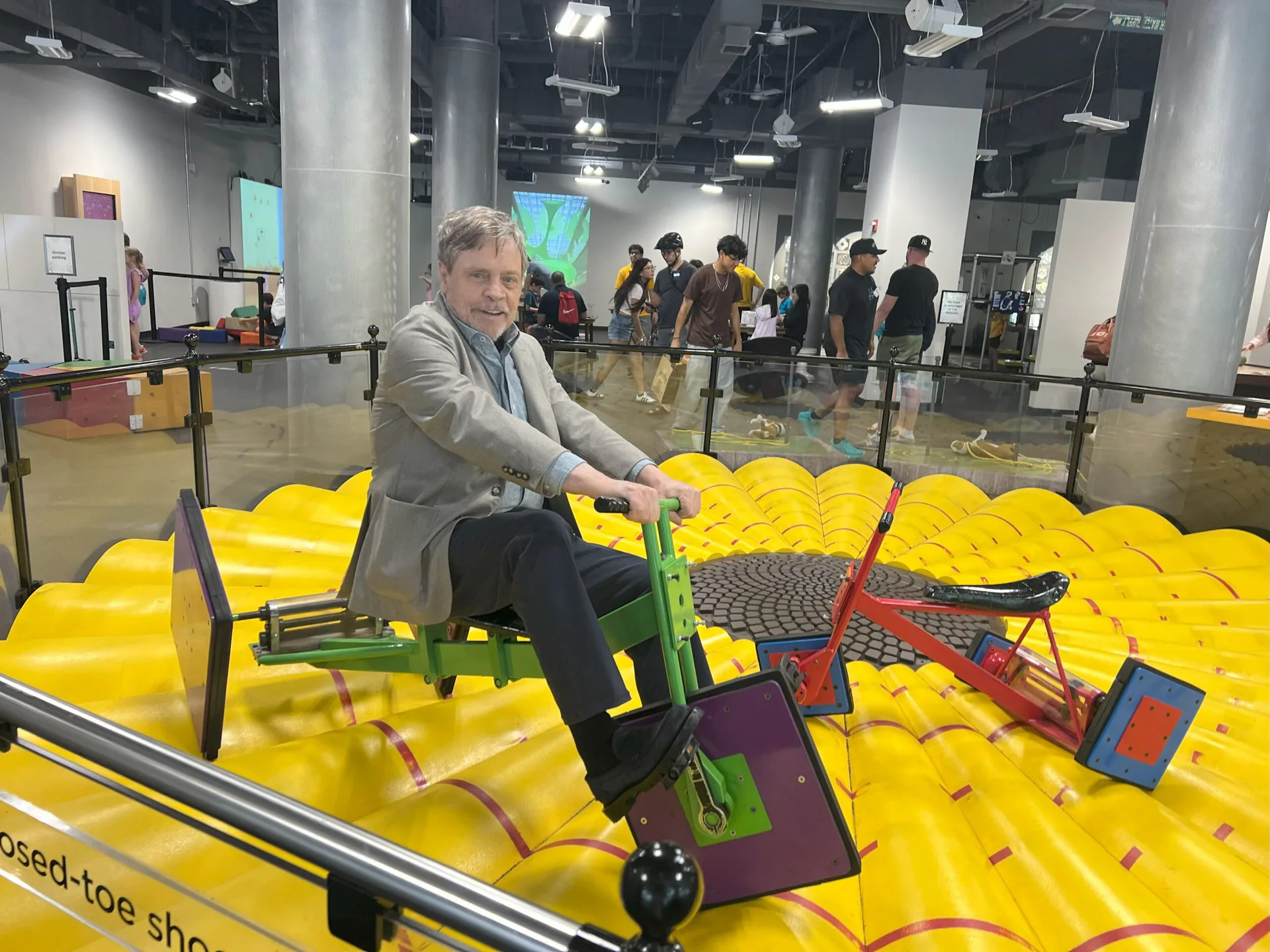 Man in gray jacket sits on green chair atop large yellow interactive floor installation at National Museum of Mathematics, with colorful objects and visitors visible in background.