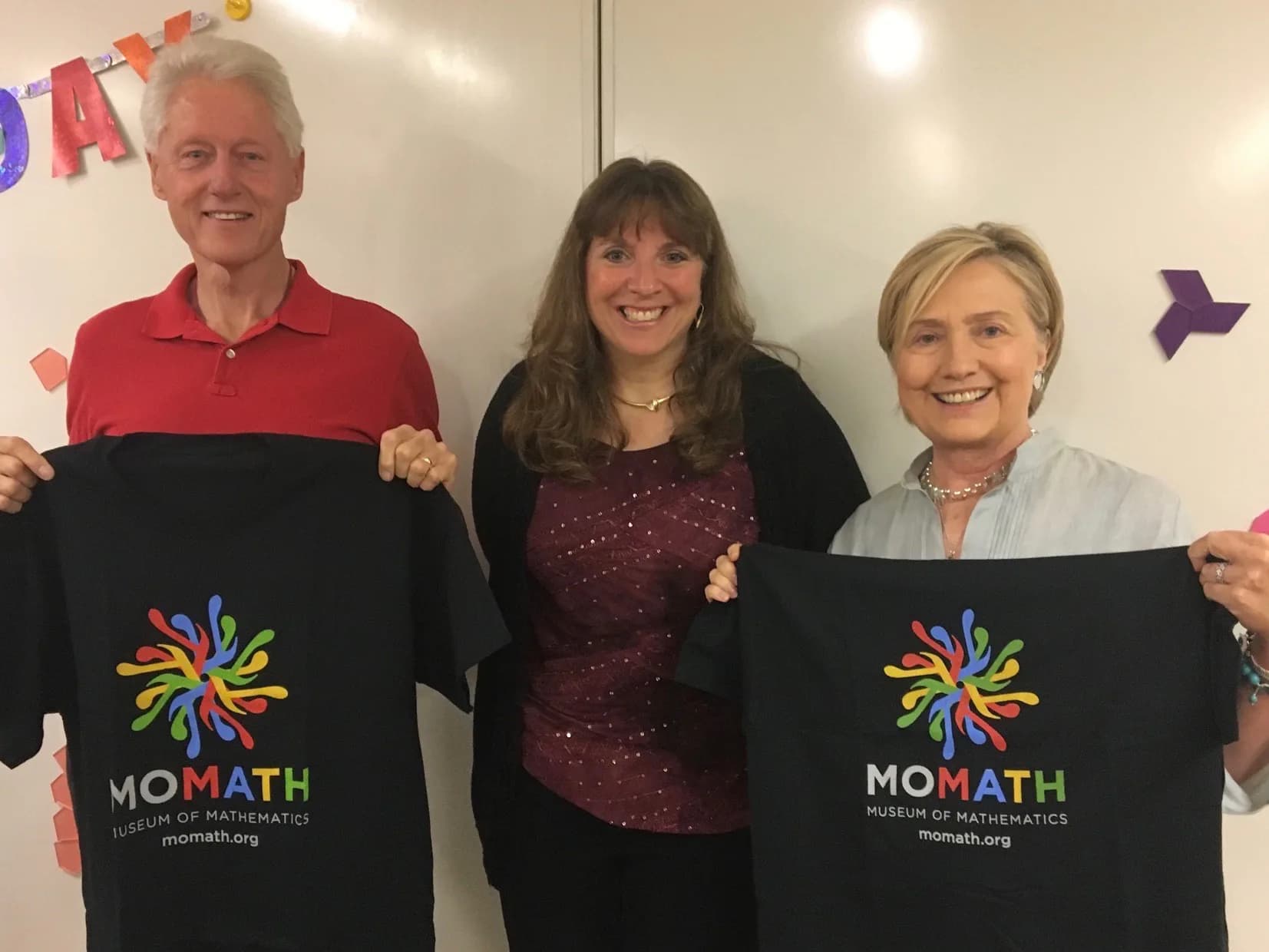 Three adults holding black MOMATH t-shirts with colorful spiral logo, smiling at camera against white wall.