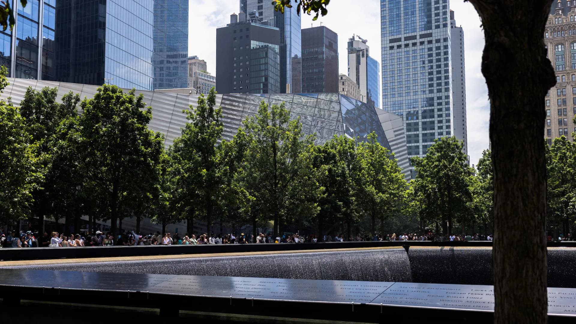 Reflecting pool at the National September 11 Memorial with trees and Manhattan skyscrapers in the background under blue sky.