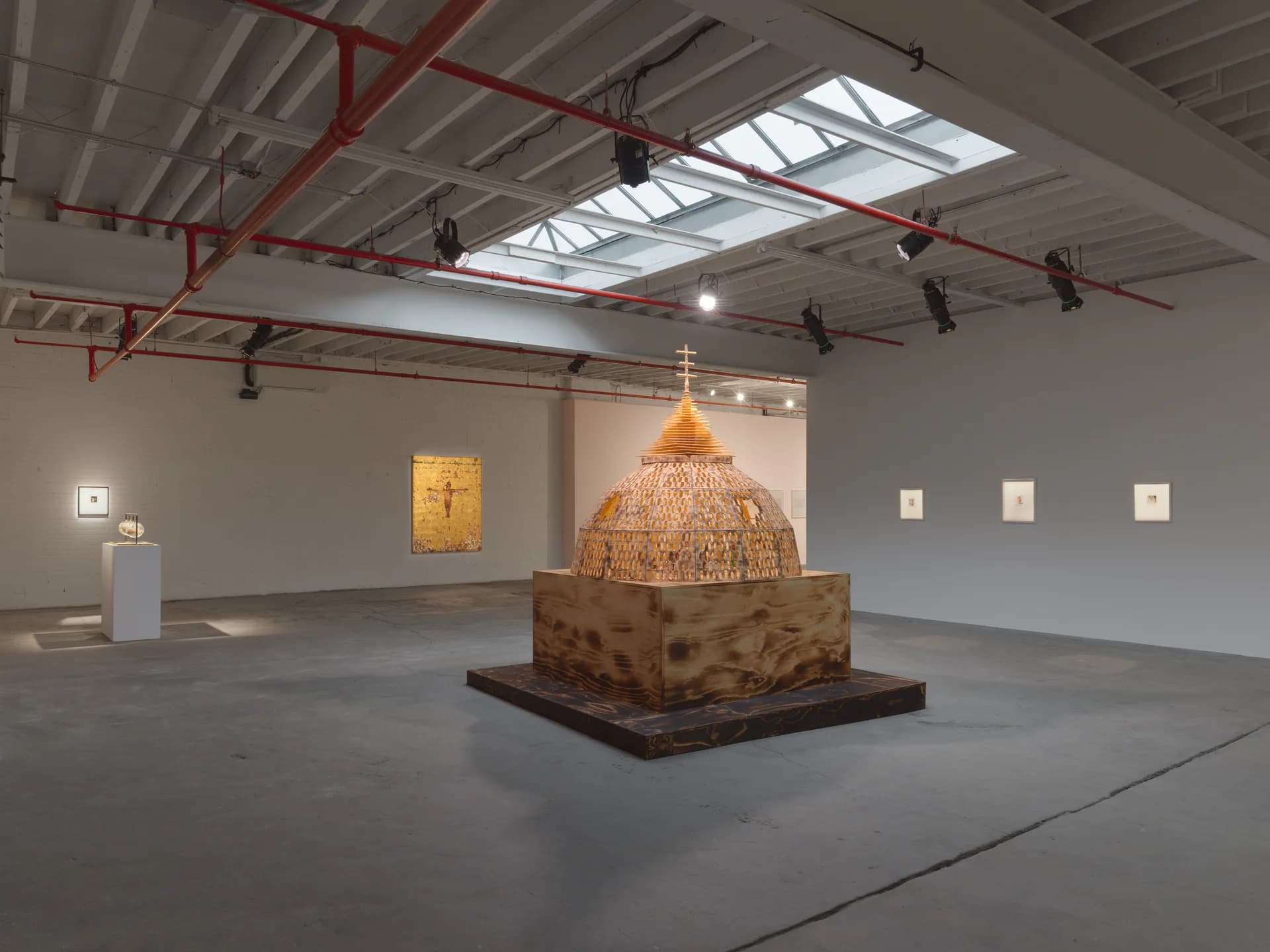 Interior gallery space with industrial ceiling, skylights, and a large bronze sculptural dome on a wooden platform in the center.