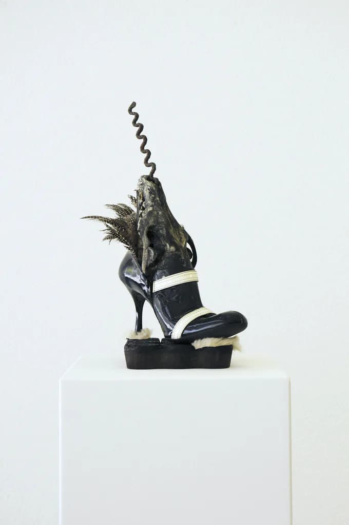 Sculptural assemblage on white plinth combining a black shoe with natural materials including twigs and feathers, with a spiral metal element extending upward.