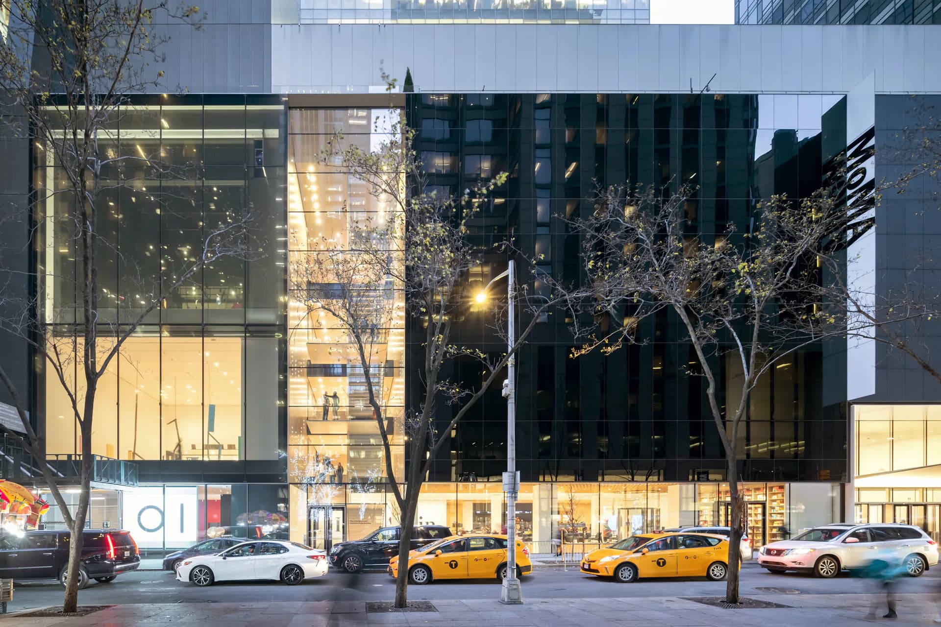 Exterior storefront of New Museum of Contemporary Art at dusk, featuring glass and dark facades with illuminated interior galleries and bare trees, yellow taxis parked on street.