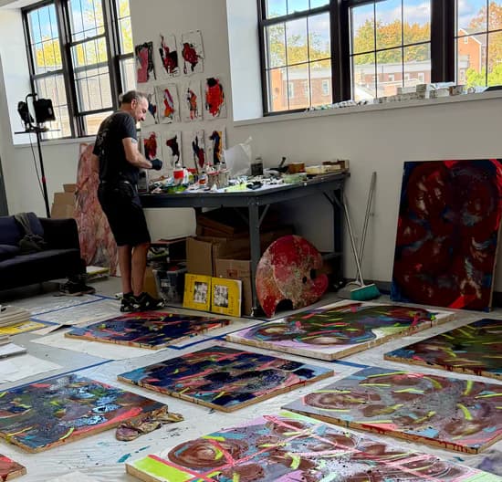 Artist working at table in bright studio with large windows; colorful abstract paintings and mixed-media works scattered on floor.