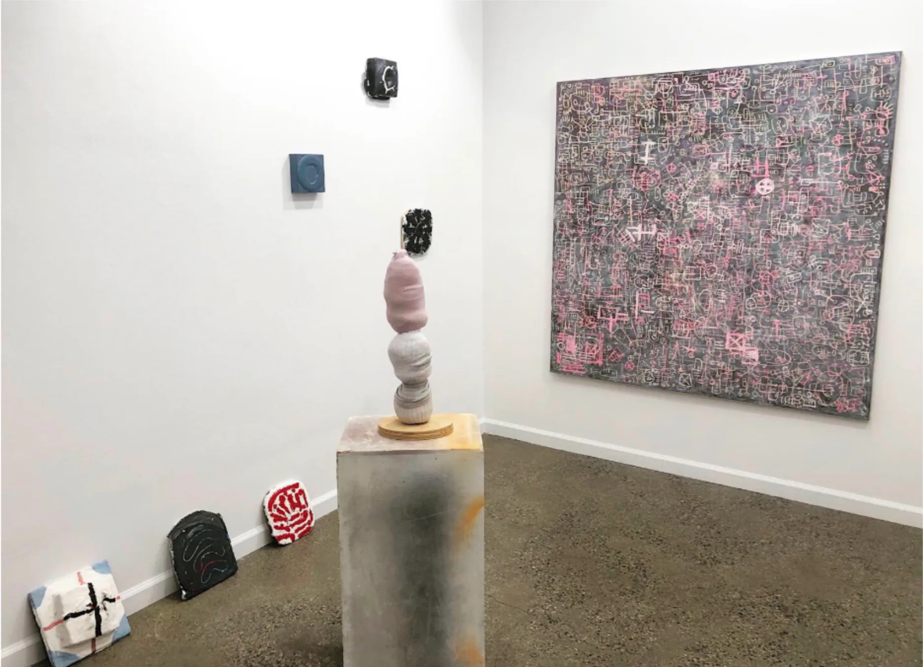 Gallery installation with stacked sculptural objects, abstract wall piece in pink and gray tones, small ceramic and found-object components.