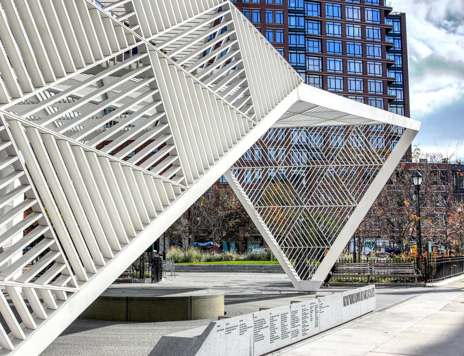White geometric sculpture with angular, ribbed triangular forms in an urban plaza, featuring cutout openings framing green trees and buildings beyond.
