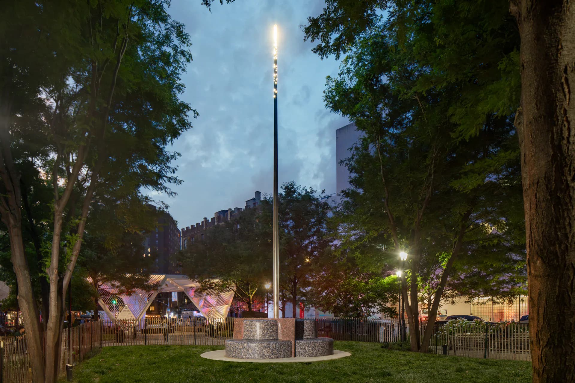 Evening photograph of an outdoor memorial site with a tall illuminated shaft, surrounded by trees, fencing, and park benches in an urban green space.
