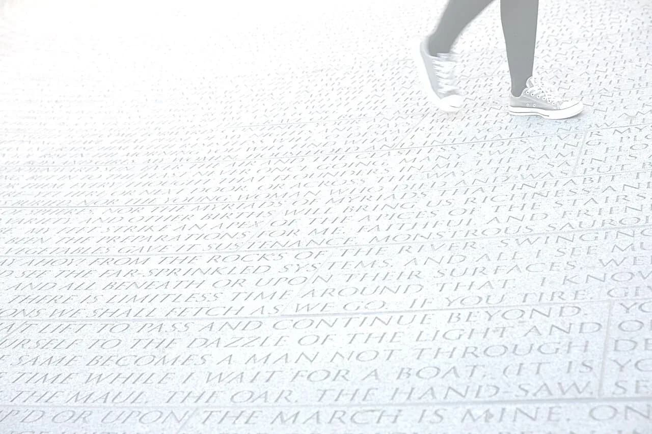 Close view of white ground surface engraved with densely packed text names; person's feet visible walking across.