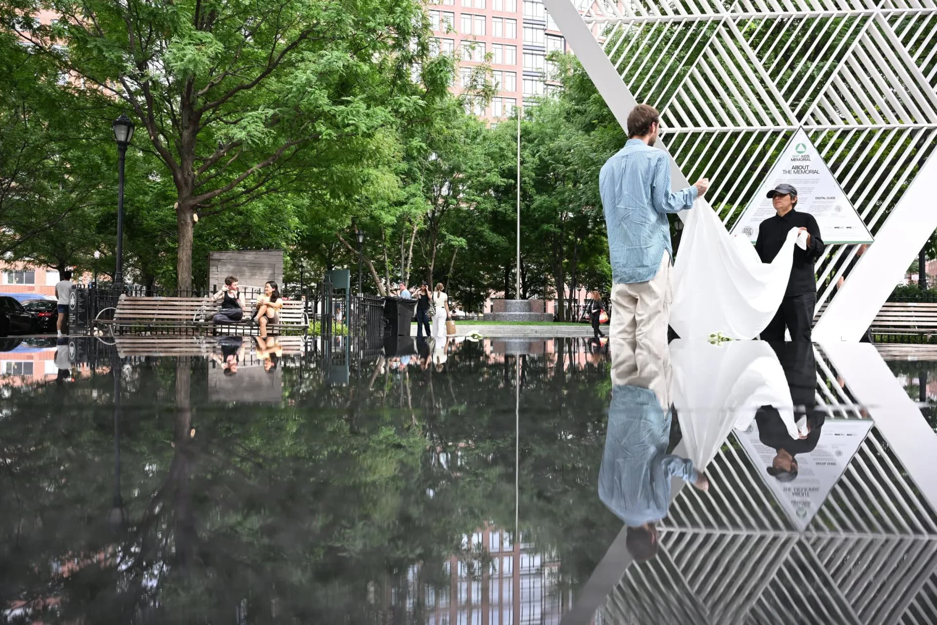 Park setting showing white geometric sculpture on right reflected in water on left, with green trees and urban buildings in background; two people present.