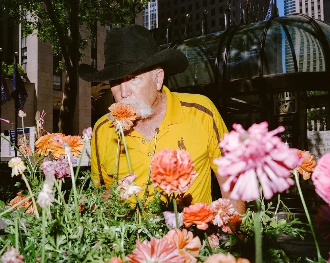 Color photograph of person in yellow jacket and wide-brimmed hat surrounded by pink and orange flowers against urban architecture backdrop.