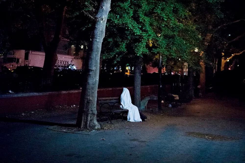 Black and white photograph of figure in white clothing seated under tree in dark urban park setting at night.