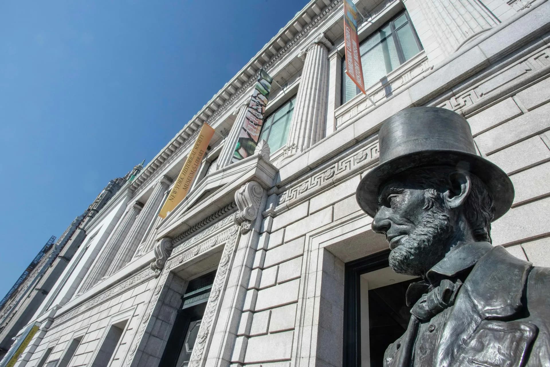 Statue of a figure wearing a top hat against a building exterior with fluted columns, stone facade, and arched doorway.