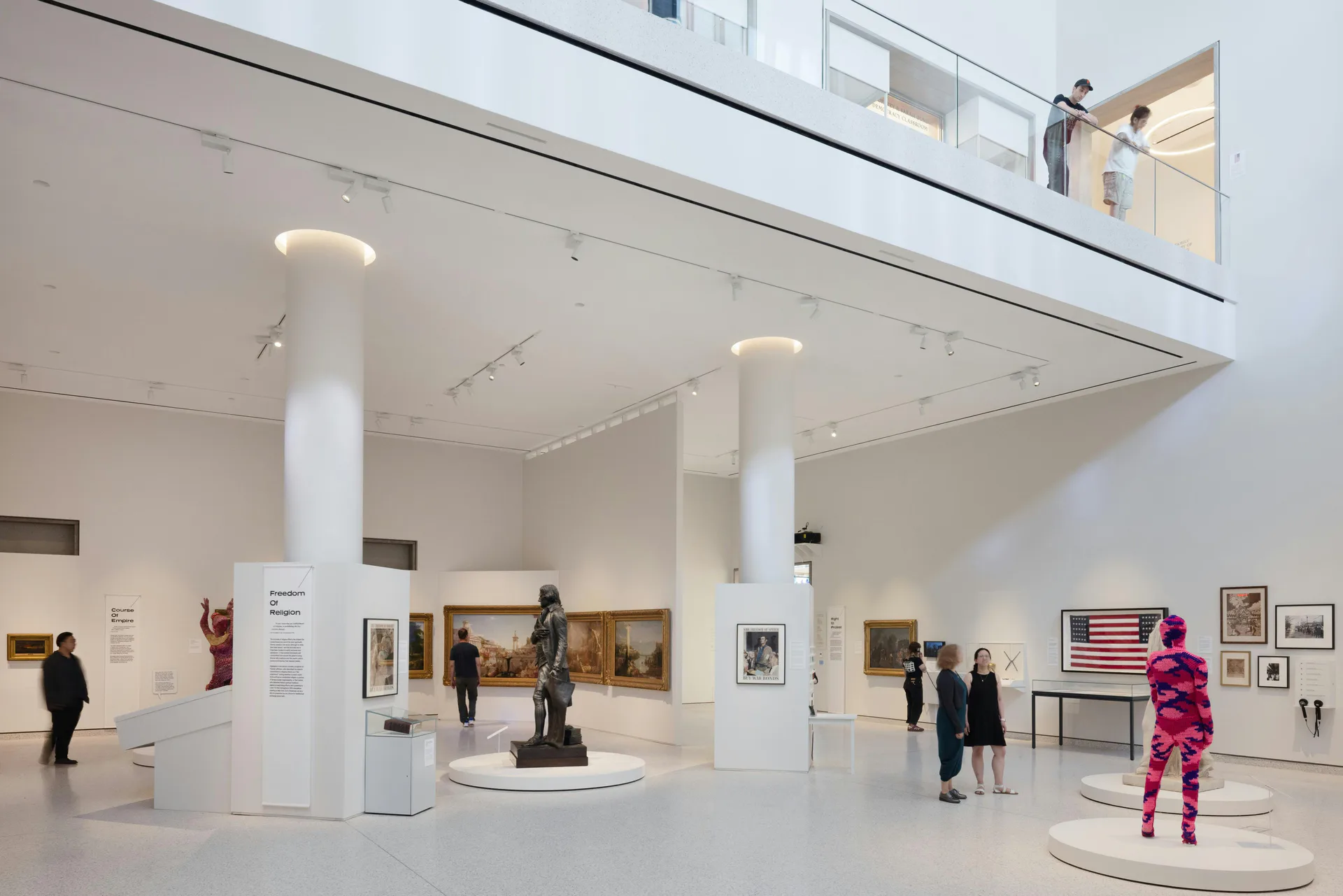 Modern white gallery interior at New York Historical Society with support columns, polished floors, sculptures and paintings displayed, visitors walking through.
