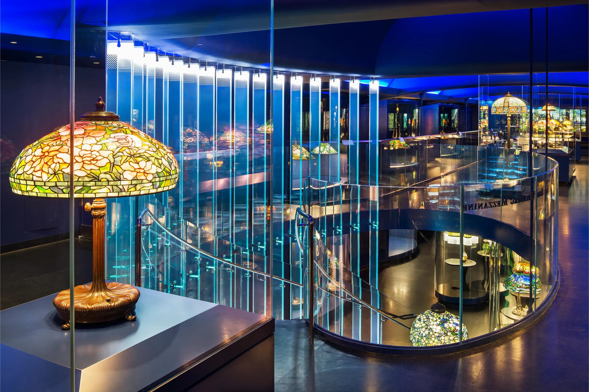 Museum gallery at dusk with glass display cases containing decorative objects, including a green glass lamp, cityscape visible through windows behind.