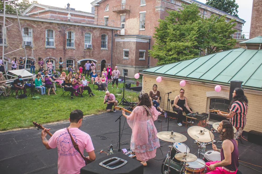 Outdoor event at Snug Harbor with live band performing on stage, brick buildings and crowds of people enjoying music.